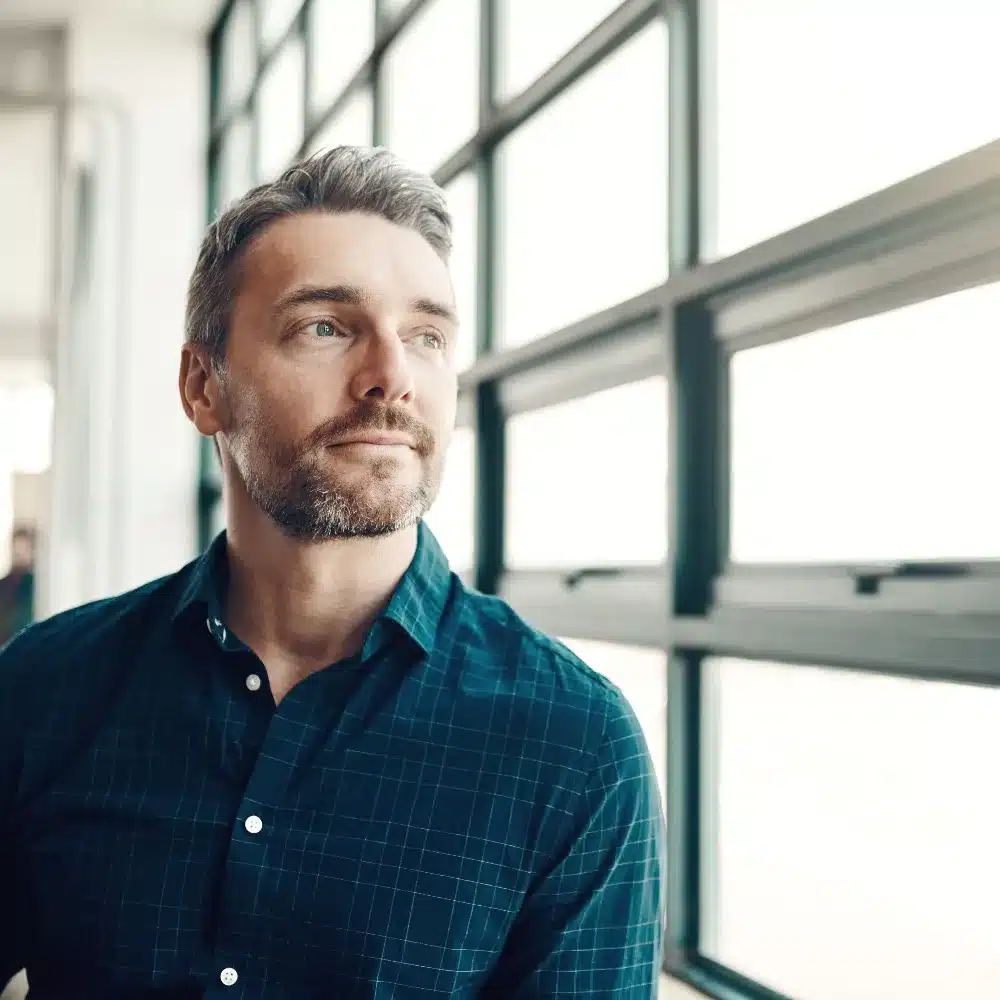 A man stands at a bright window, deep in thought